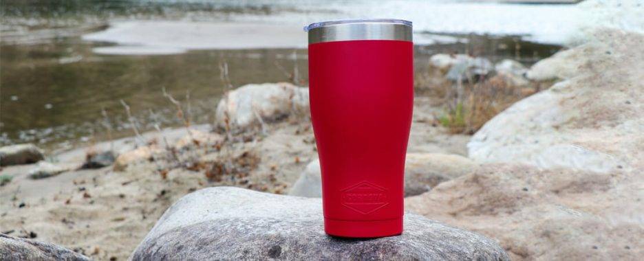 Red Cordova tumbler sitting on a rock wiht a river in the background